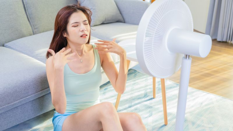 junge frau sitzt schweißgebadet vor einem ventilator im wohnzimmer und lässt sich kühlen