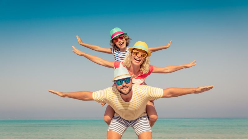 papa, mama und kind im sommerlichen outfit mit strohut und sonnenbrille steehn übereinander mit ausgestreckten armen am meeresstrand