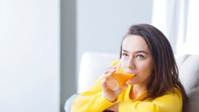 junge sportliche frau sitzt im sofa und trinkt einen orange farbenen curcuma smoothie
